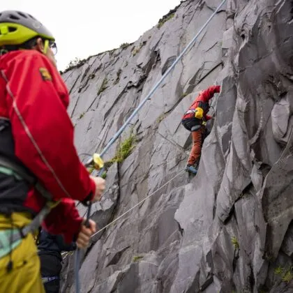 Person climbing a ruggid cliff being supported by someone stood on the ground wearing a red jacket and yellow helmet