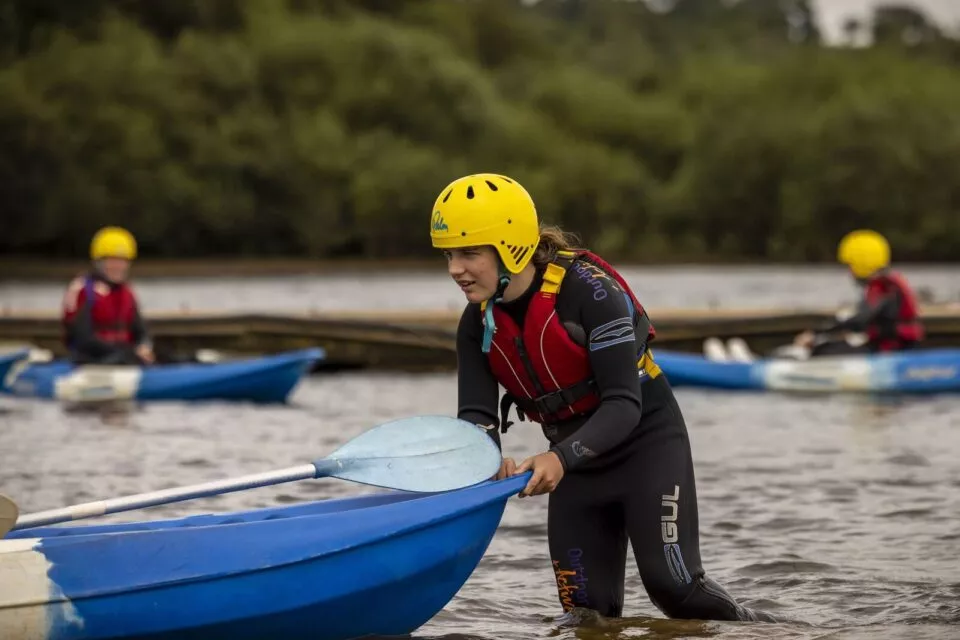 cadet in wetsuit and yellow helmet standing in a lake pushing a dark blue boat with a light blue paddle balanced on top