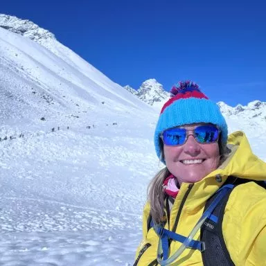 Female with sunglasses and beanie poses with snowy mountain peak behind her