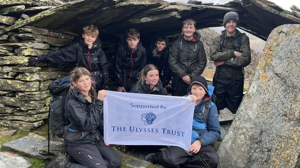 Cadets crouching under a stone monument, holding the Ulysses Trust flag